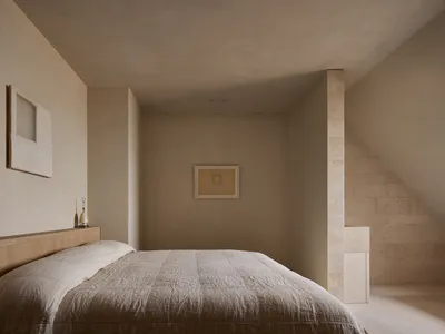 極簡臥室室內空間，大床鋪著質感米白色亞麻寢具，木質床頭板層架上擺放兩盞陶瓷檯燈，洞石牆面、斜頂天花板，牆上掛有抽象藝術畫框。