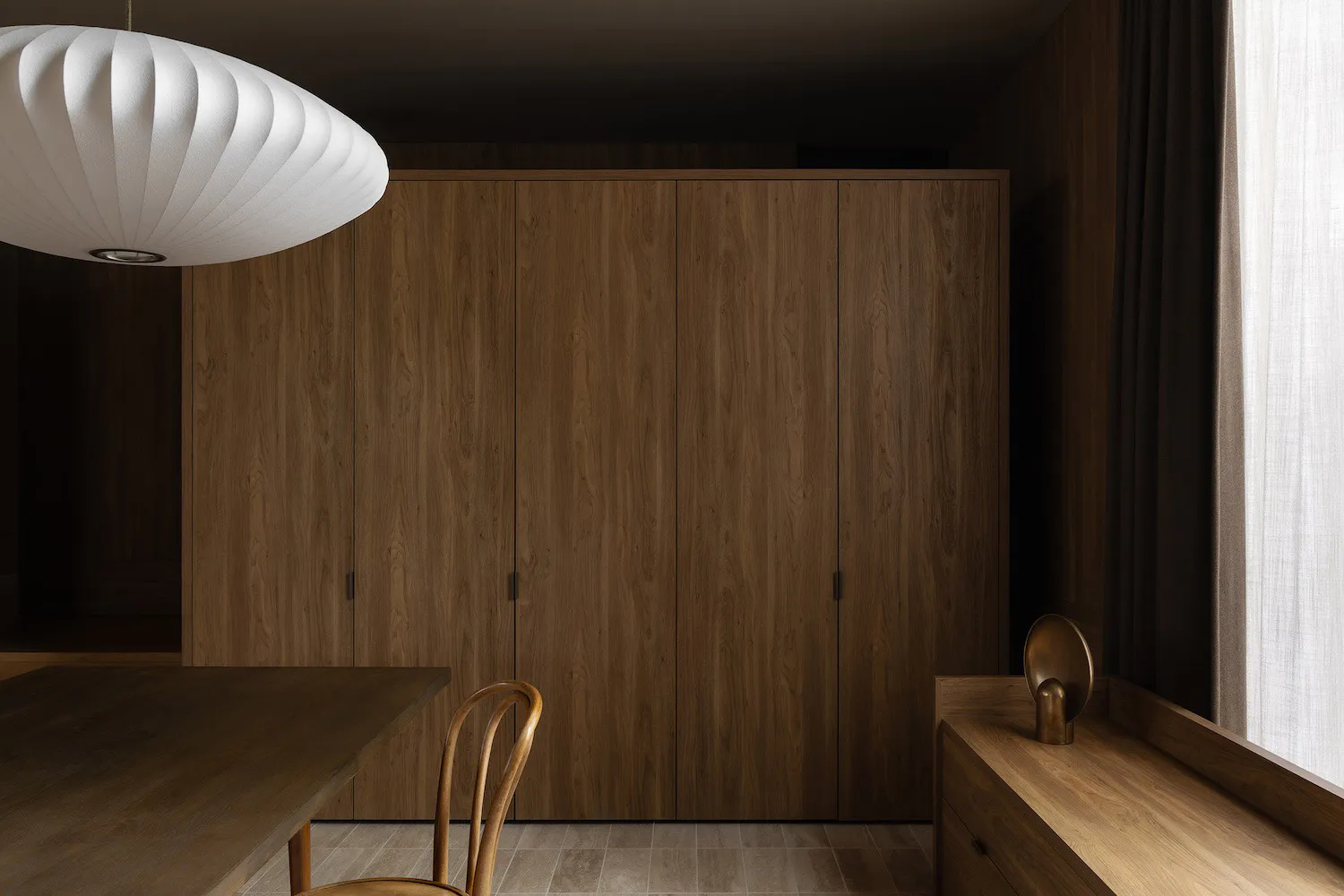 Modern minimalist dining area featuring a large walnut wood wardrobe, wooden dining table and chair, sculptural white pendant lamp, and a window seat with brass decor.