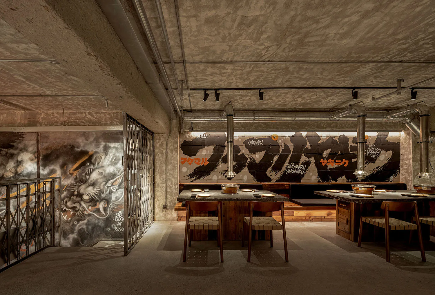 Interior view of an industrial-style Japanese yakiniku restaurant featuring exposed concrete walls, large dragon graffiti mural, stainless steel exhaust hoods, and wooden dining tables with woven chairs.