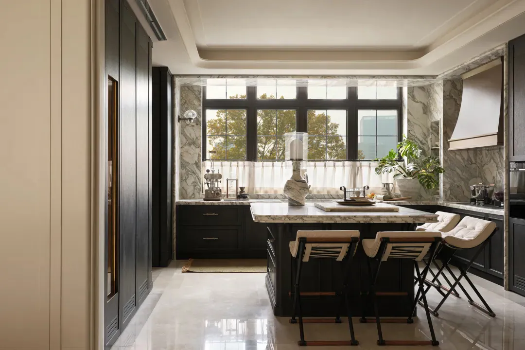 Modern luxury kitchen featuring a large marble island with three bar stools, dark navy cabinetry, marble backsplash, and a wall of tall black-framed windows allowing abundant natural light.