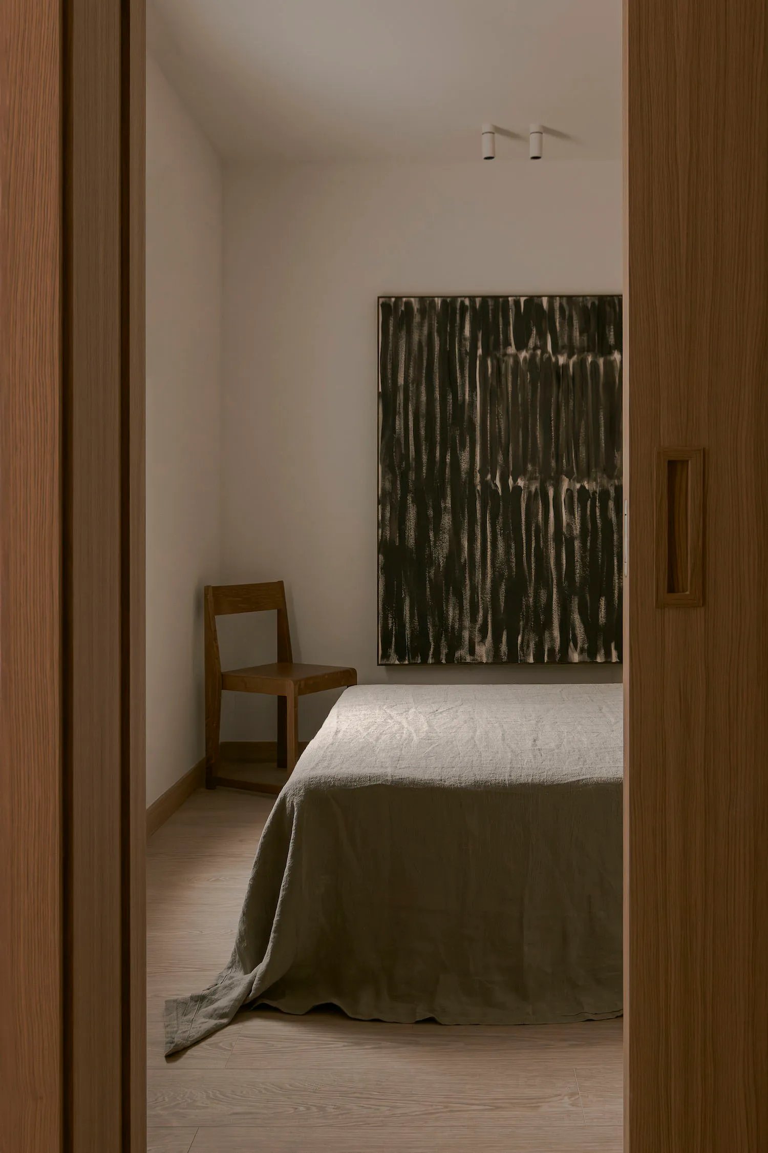 View into a minimalist bedroom with light oak wood elements, a wooden chair, a bed with rumpled neutral linen bedding, and a large black and white abstract painting on the wall.