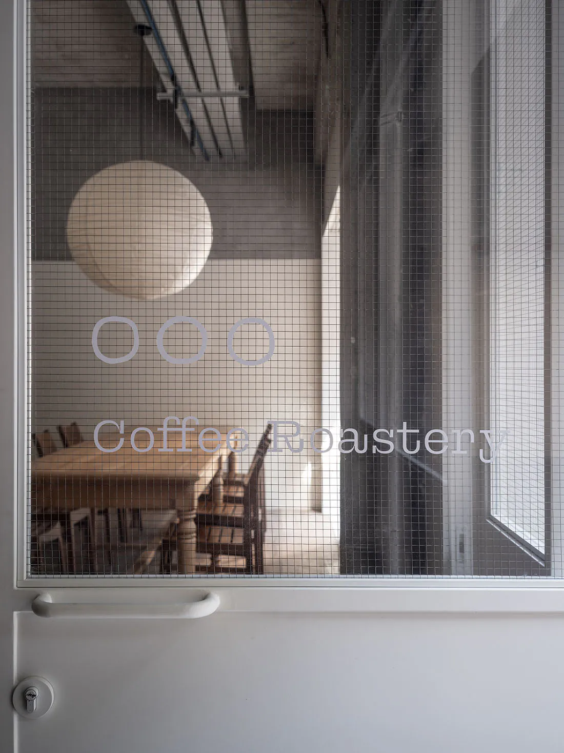 View through a white-framed wire mesh door with handle and lock into a coffee roastery interior featuring a large round pendant lamp, wooden communal table, and exposed ceiling pipes.
