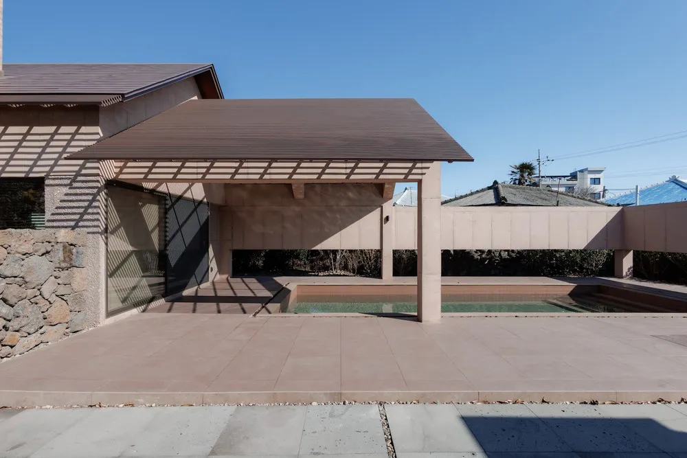 Modern minimalist concrete courtyard with a shallow reflecting pool, wooden pergola casting linear shadows, and a stone wall on the left under a clear blue sky.