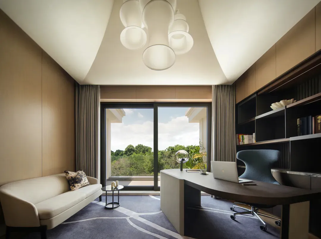 Modern minimalist home office featuring a curved walnut desk, ergonomic blue office chair, cream sofa, sculptural white pendant light fixture, and large glass doors opening to a green forest view.