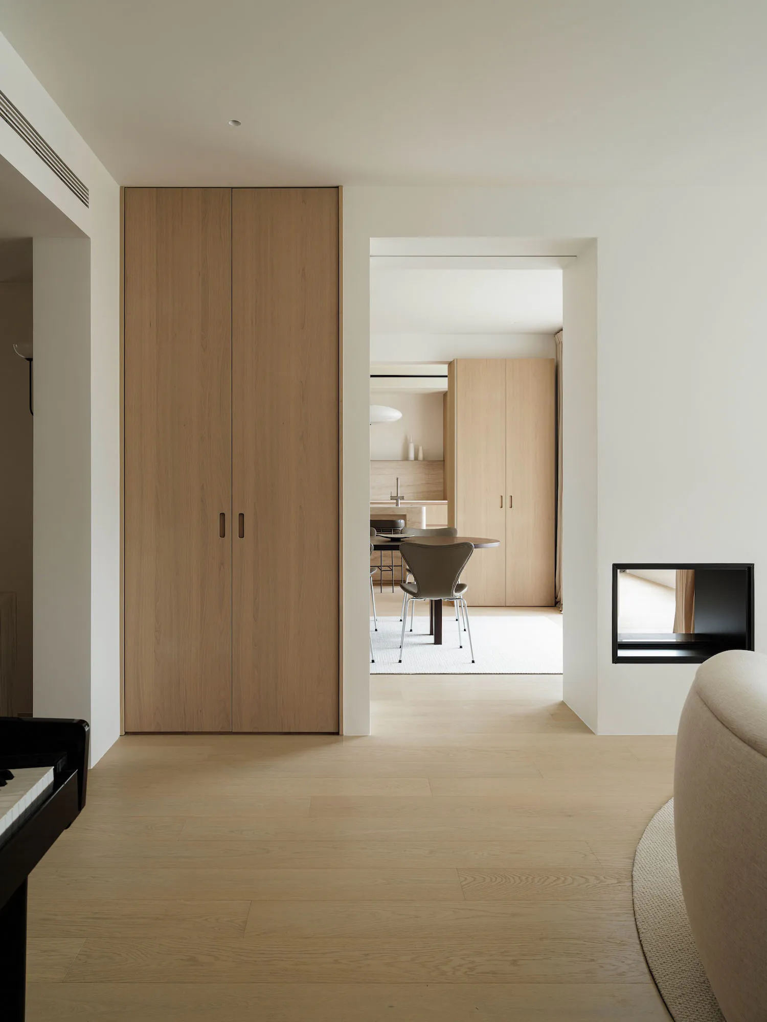 View from a minimalist living room through a wide opening into a light oak dining area with round table and grey chairs, featuring built-in wood storage and a black fireplace niche.