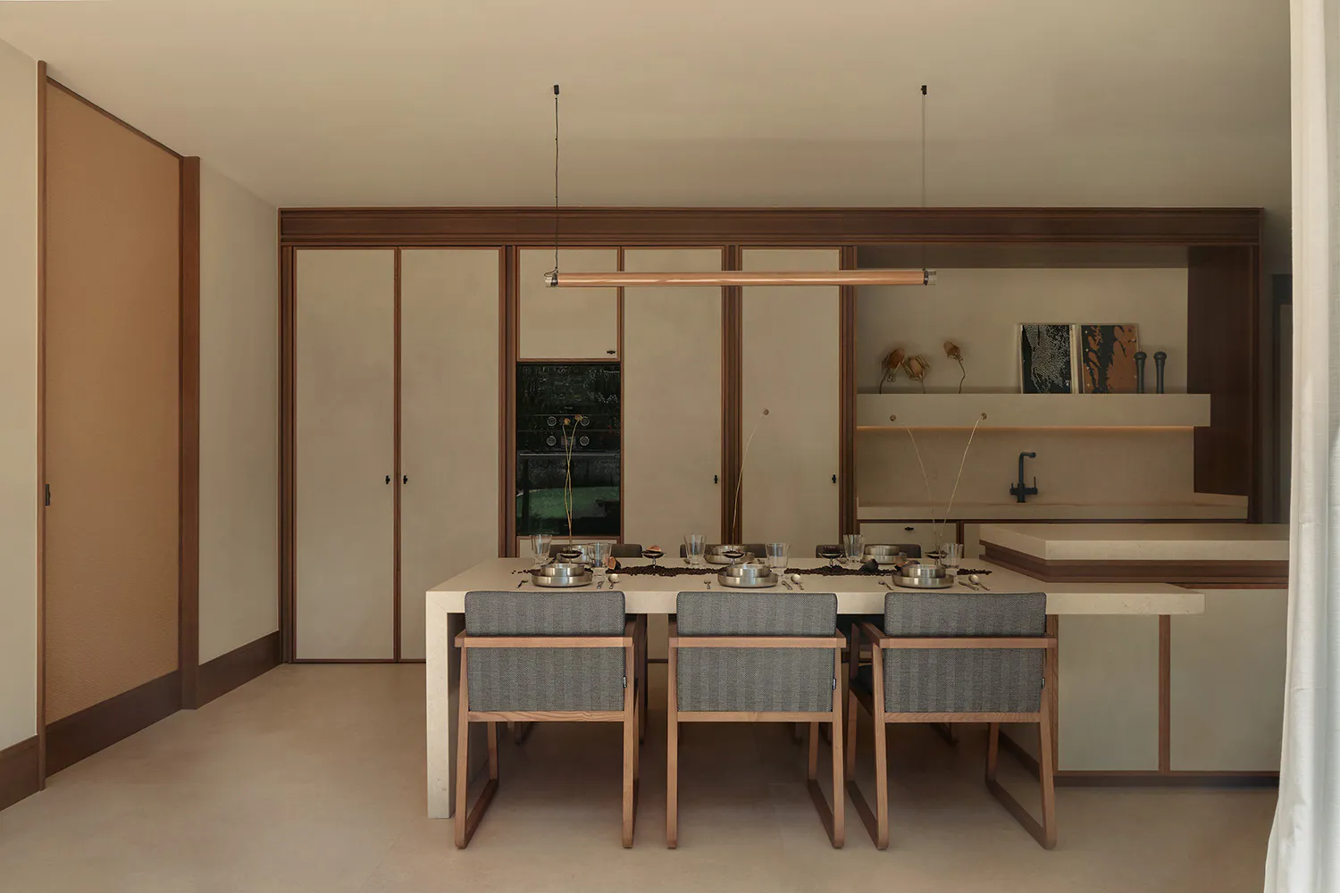 Modern minimalist dining room featuring a long concrete dining table with six wooden and fabric chairs, a linear copper pendant light, and a walnut wood kitchen cabinetry with open shelves and integrated sink in the background.