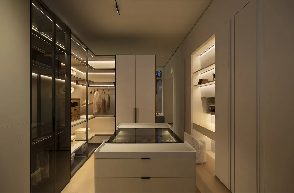 Modern luxury walk-in closet with white matte cabinetry, central glass island, built-in LED lighting, and organized clothing storage.