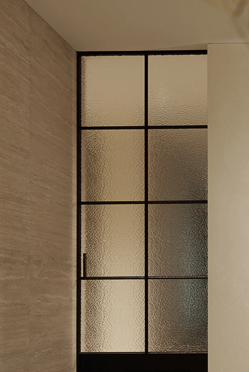 Modern interior door with black metal grid frame and textured glass panels set between a concrete wall and a white wall.
