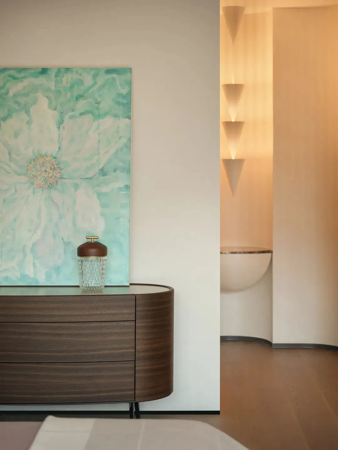 Modern minimalist entryway featuring a curved dark walnut console table with three drawers, a crystal and wood lidded jar, and a large abstract turquoise and white peony painting on a white wall. To the right is a curved niche with a modern vessel sink and a vertical tiered pendant light fixture emitting warm illumination.