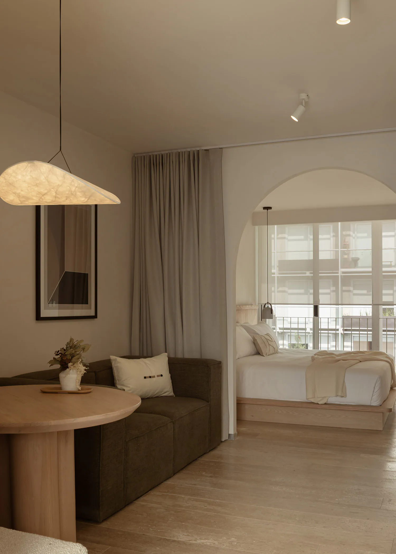 Modern minimalist interior featuring a living area with brown modular sofa, round oak table, and large arched opening revealing a bedroom with platform bed and large windows.