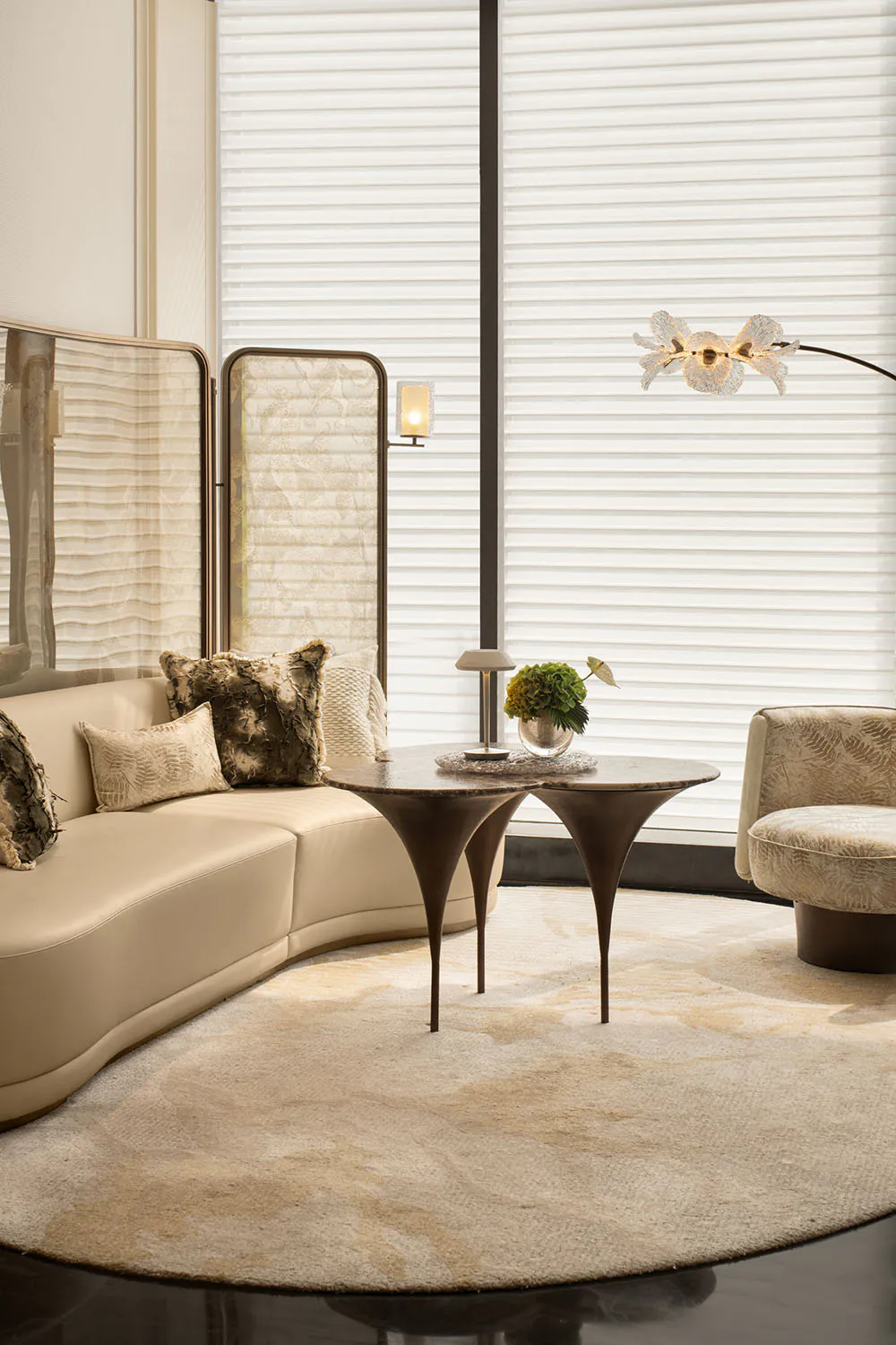 Modern luxury living room with a curved cream leather sofa, two dark marble nesting coffee tables, a patterned armchair, and an orchid floor lamp in front of large windows with horizontal blinds.