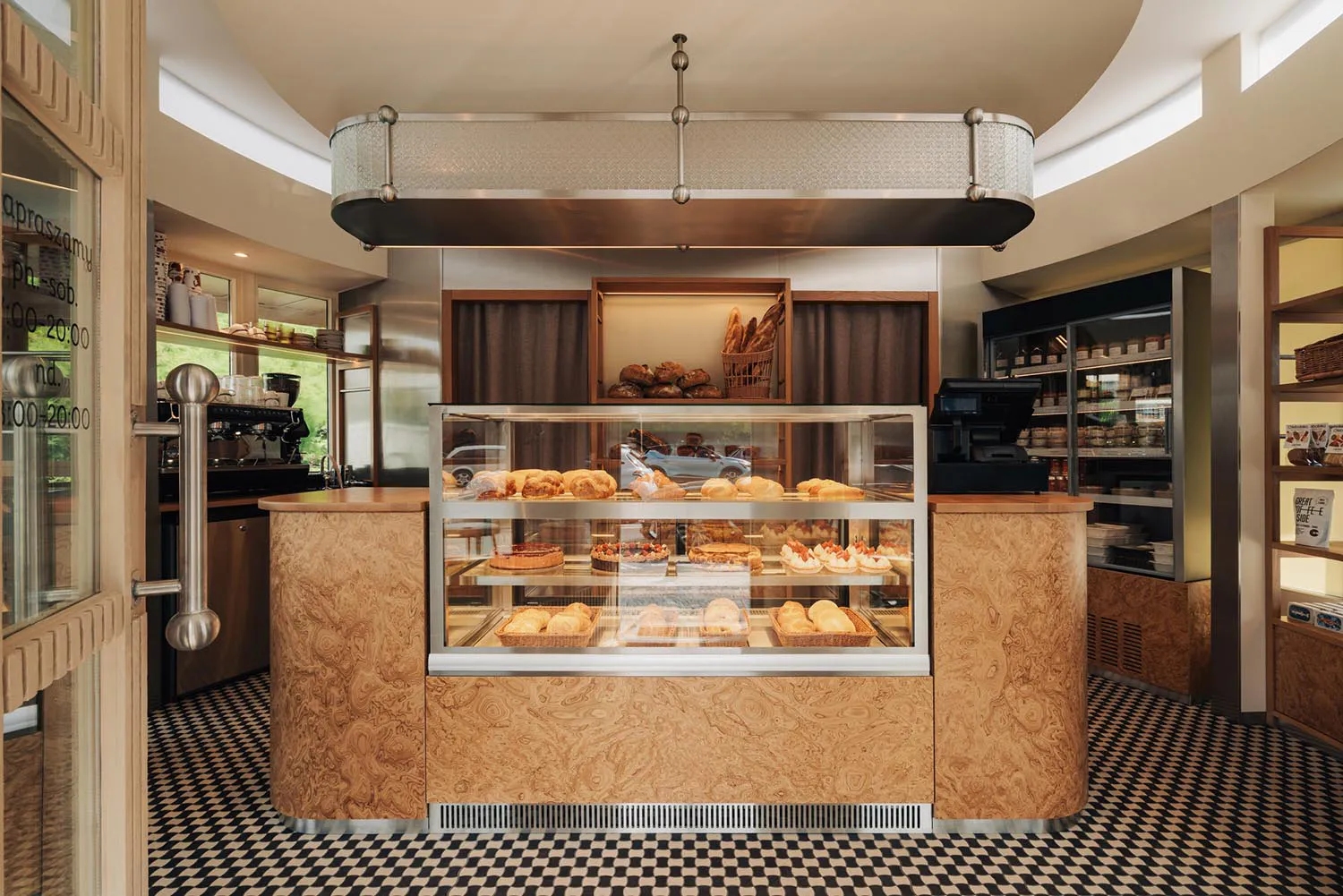 Interior view of a stylish bakery featuring a large curved burl wood display counter with glass pastry case, black and white checkerboard floor, and pendant lighting.