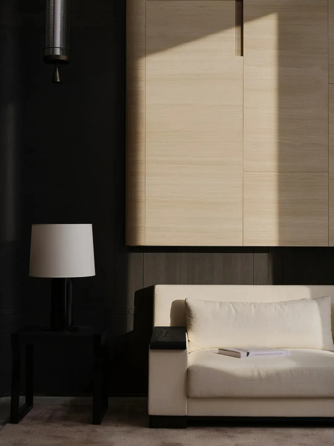 A contemporary living room corner featuring a cream-colored sofa with a book on the seat, a black side table with a white table lamp, and large light wood wall cabinetry with strong diagonal sunlight casting shadows, against a dark wall with a hanging pendant light.