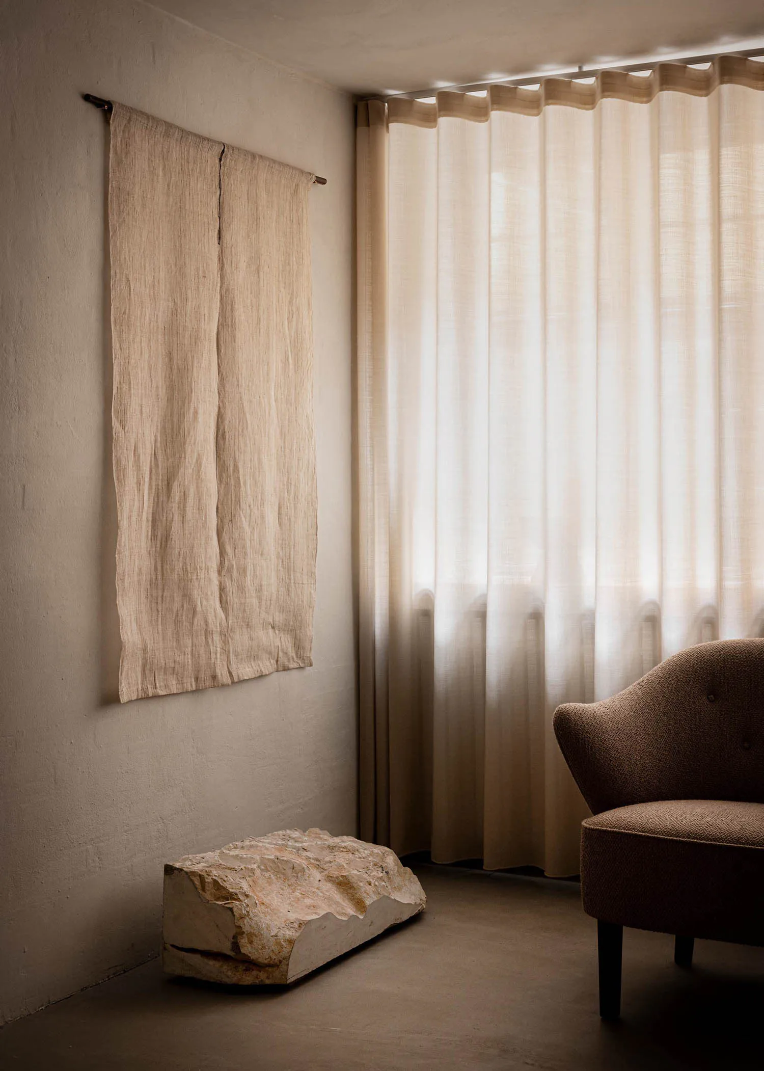 A minimalist interior corner featuring two hanging natural linen panels on a white wall, a large rough-hewn stone block on the floor used as a bench, sheer linen curtains diffusing daylight, and a brown bouclé upholstered armchair in the foreground.