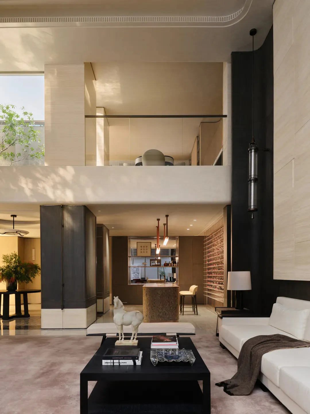 Modern luxury double-height living room with travertine stone walls, white sectional sofa, black coffee table, and upper mezzanine level overlooking the space.