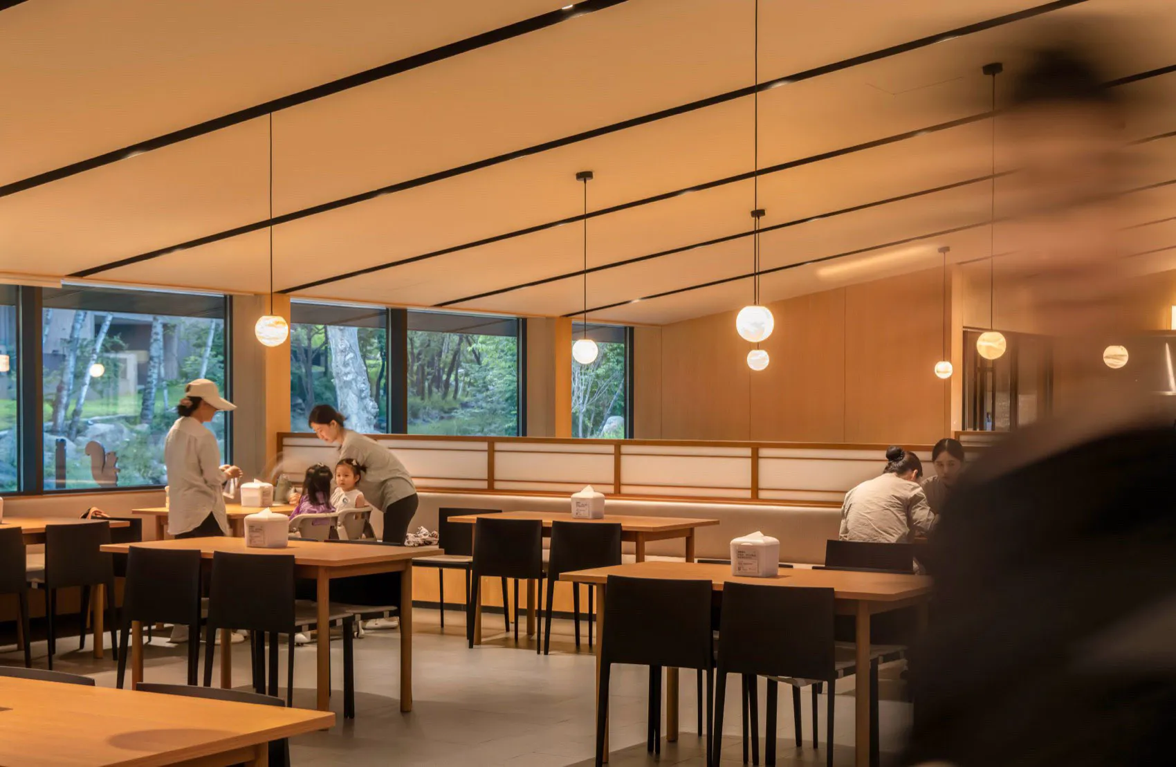 Modern minimalist family restaurant interior featuring light wood tables, black chairs, spherical pendant lights, and large windows overlooking a forest.