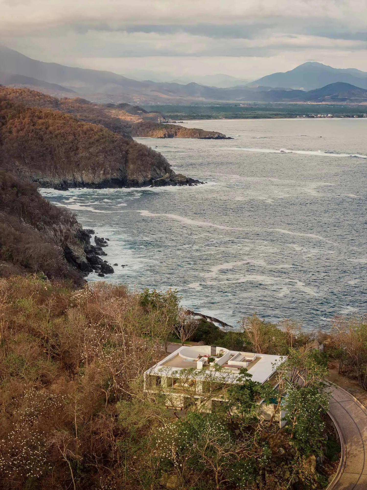 Modern concrete villa built into a forested cliffside overlooking the Pacific Ocean with distant mountains and coastline visible at sunset.