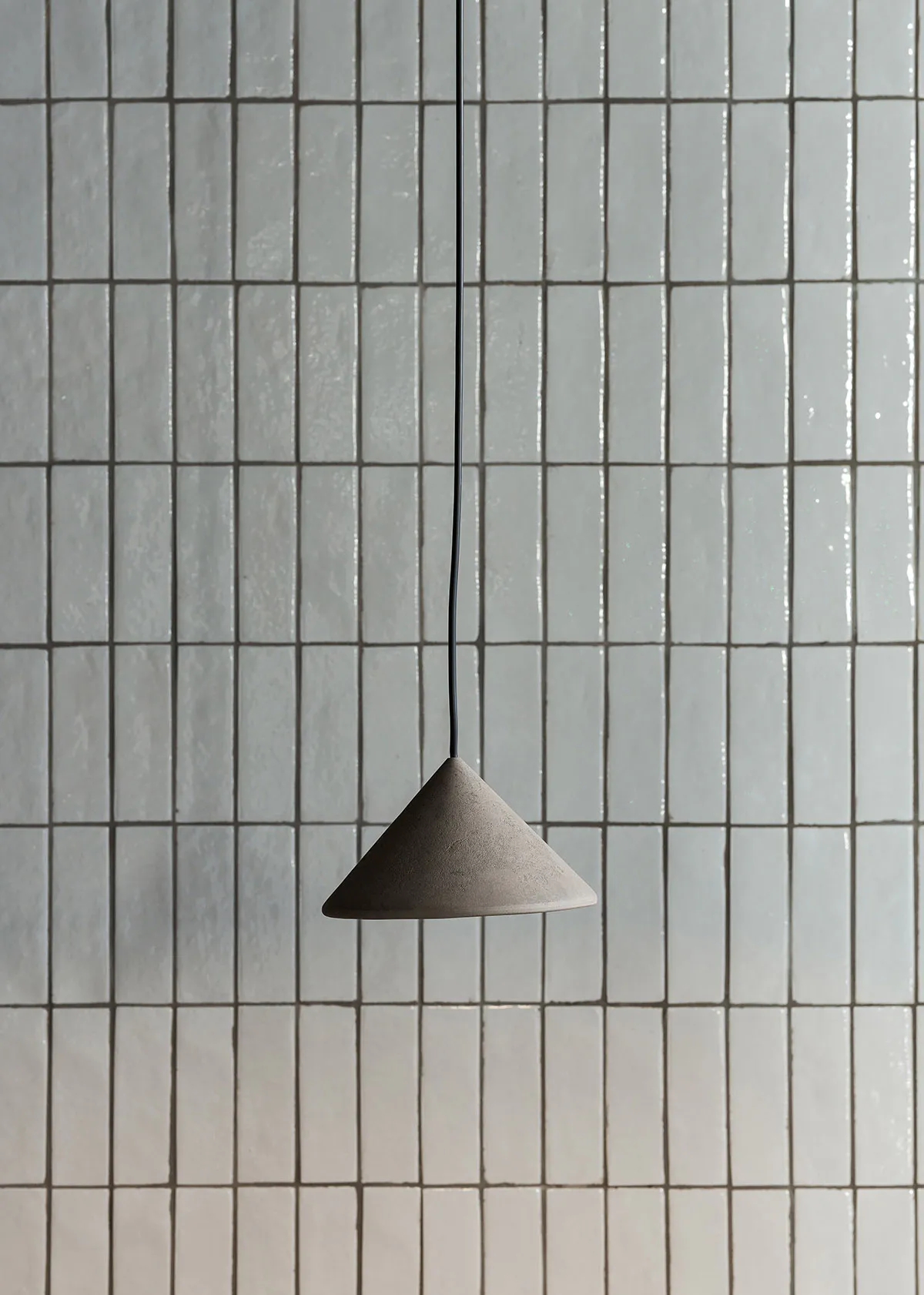 A minimalist concrete pendant light with a cone shape hanging from a black cord against a background of light grey rectangular wall tiles.