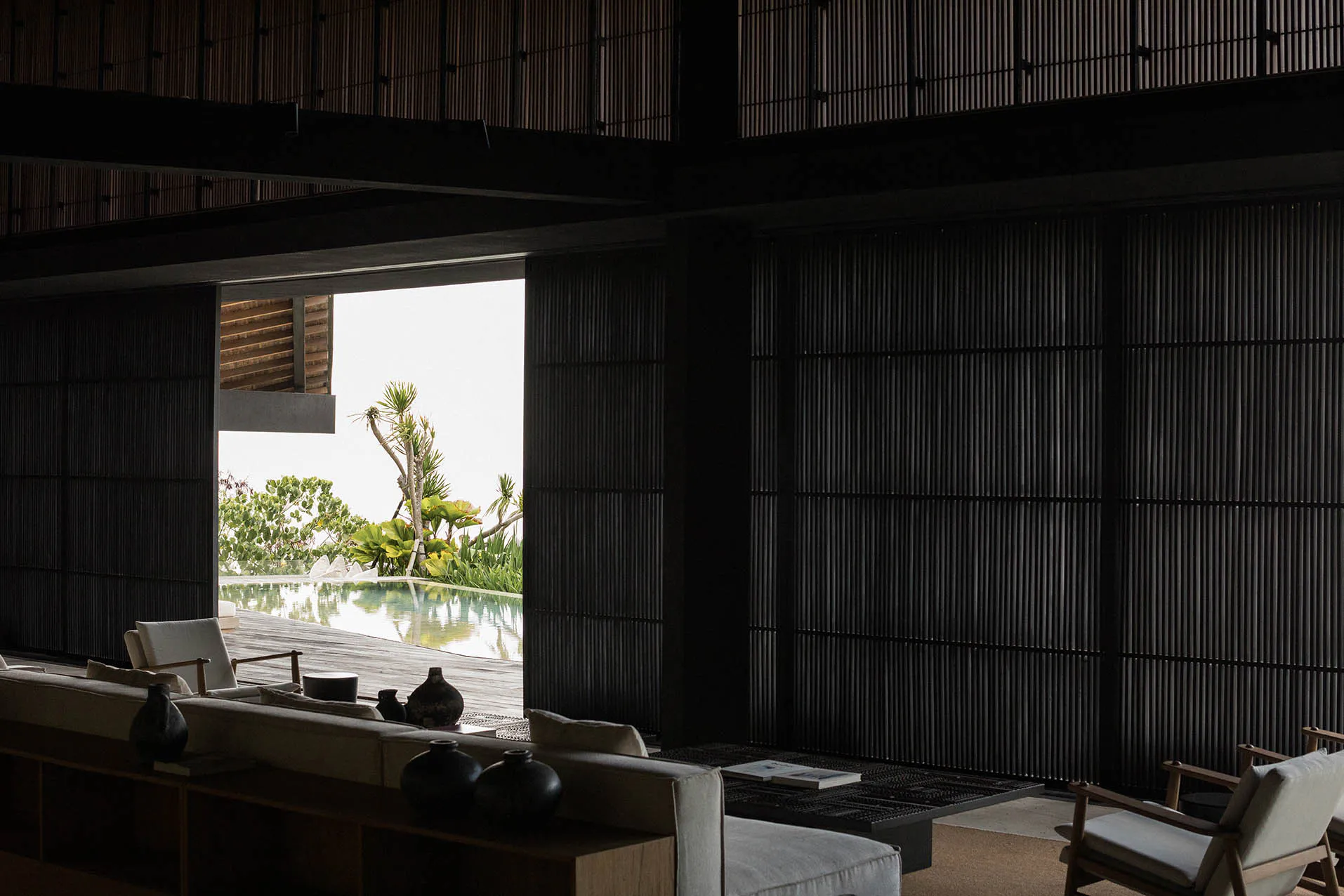 Modern tropical living room interior with dark vertical wood slat walls, light neutral sofas, wooden armchairs, black ceramic vases, and an open view to an infinity pool surrounded by tropical plants.