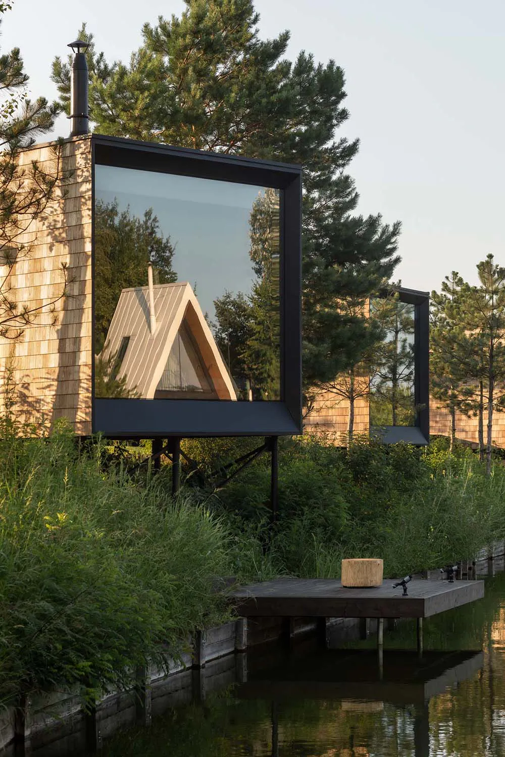 Modern wooden cabin with large black mirror installation reflecting another A-frame cabin among pine trees, situated over a calm lake with a wooden dock in the foreground.