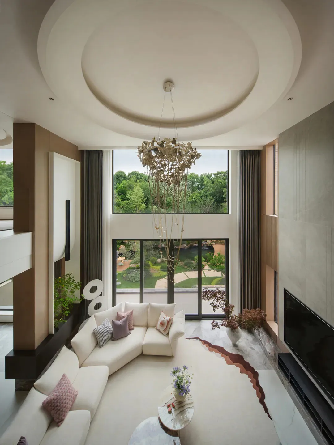 Modern luxury living room with double-height circular ceiling, large sculptural metallic chandelier, curved white sectional sofa, and marble floor with copper-colored inlay.