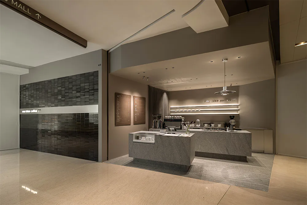 Modern minimalist cafe interior with grey stone counter, marble service island, dark tiled feature wall, and warm neutral color scheme inside a shopping mall.