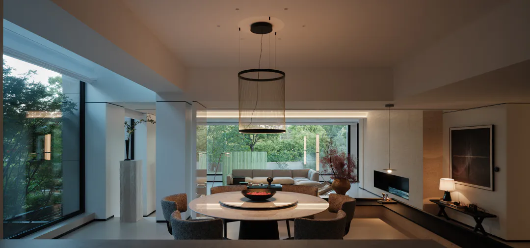 Modern minimalist dining room featuring a large round white marble table surrounded by six textured grey upholstered chairs, a tall sheer cylindrical pendant light, and large glass doors opening to a lush green garden.