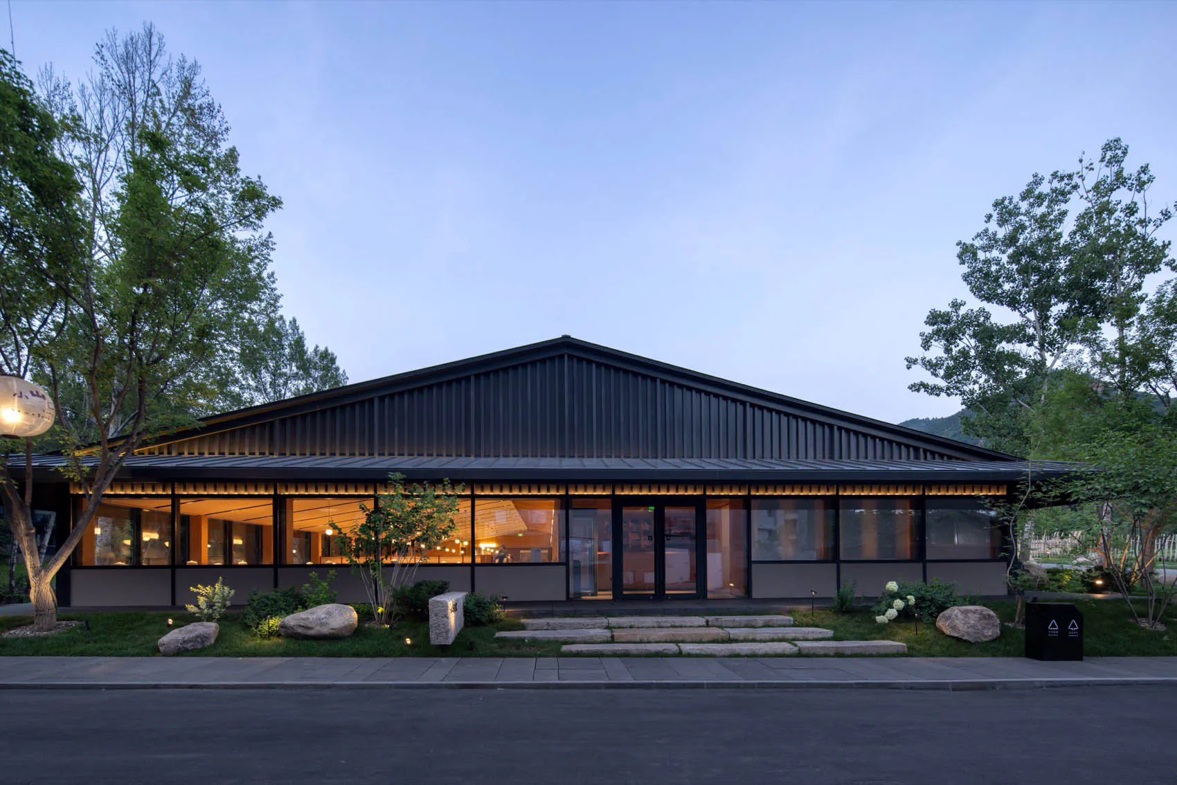 Modern single-story house with black corrugated metal roof and large glass windows glowing with warm light at twilight, surrounded by landscaped garden and trees.