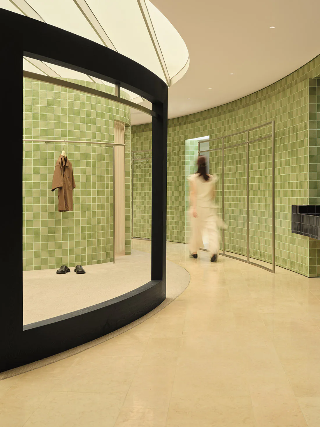 Modern boutique interior featuring curved black architectural frame, sage green ceramic tile walls, minimalist clothing display, and a blurred figure walking through the space.