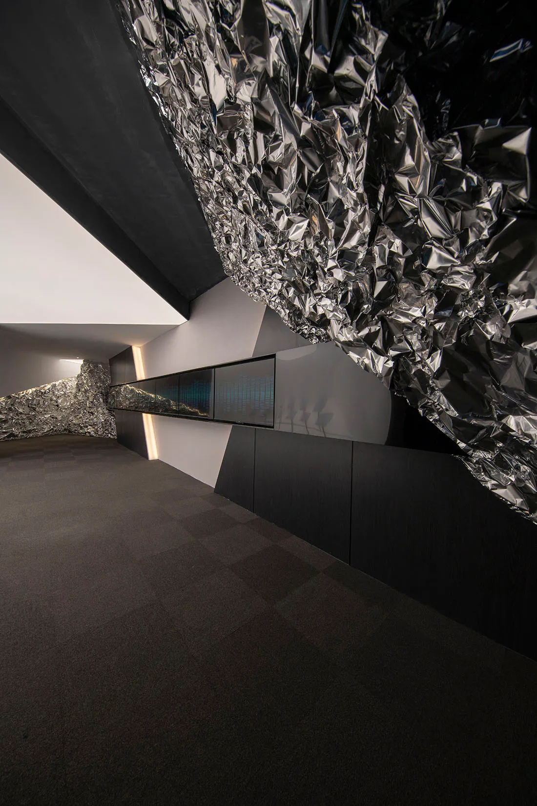 Modern art gallery interior featuring a large sculptural installation of crumpled reflective metal suspended from the ceiling, dark tiled floor, and digital display screens along the wall.