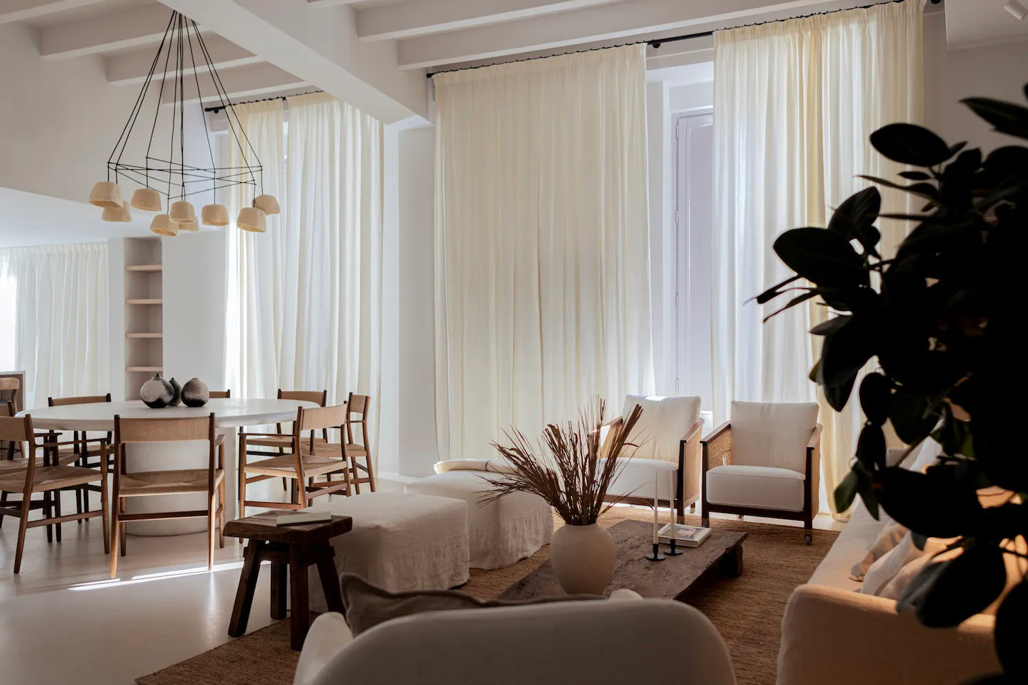 Bright open-plan living and dining room featuring a round white dining table with woven rattan chairs, a black geometric chandelier, two white armchairs with wooden frames, a low wooden coffee table, and large windows with sheer white curtains allowing abundant natural light.
