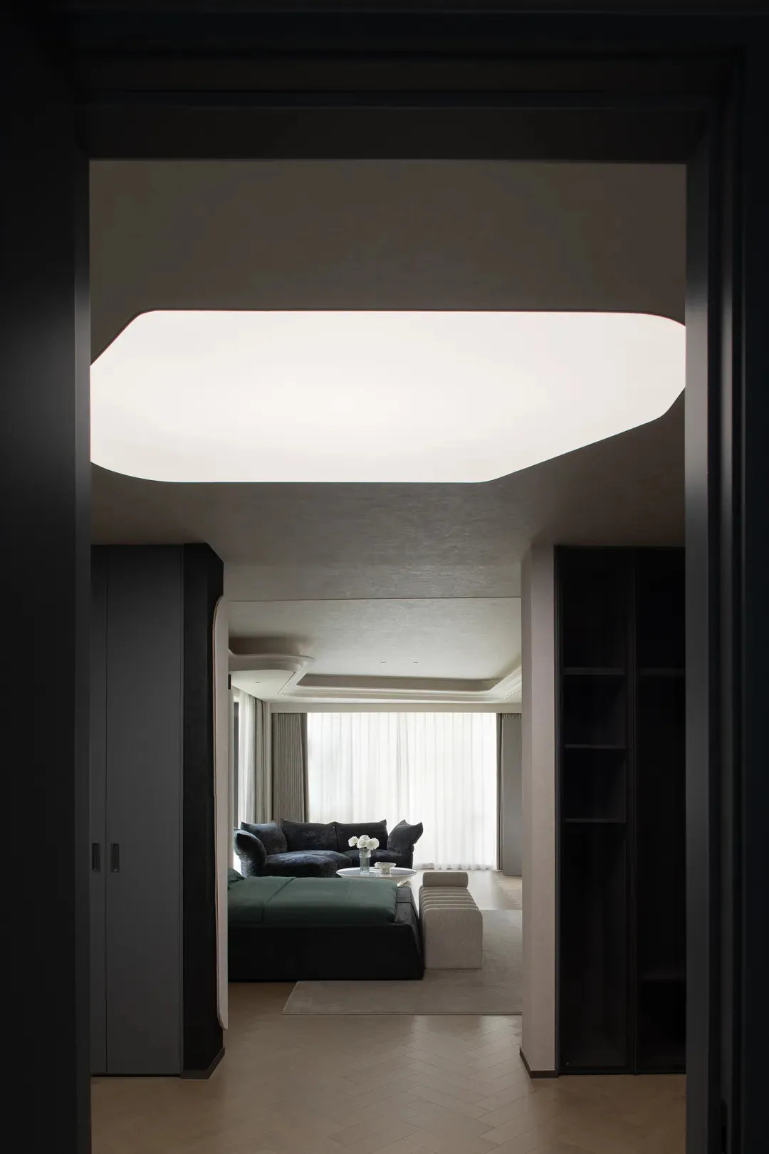 View from a dark doorway into a modern living room featuring a large sculptural ceiling light, dark green modular sofa, and light herringbone wood flooring.