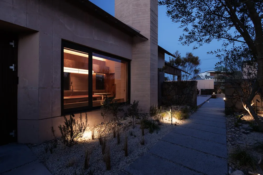 Exterior view at dusk of a modern concrete house with large black-framed windows showing a warmly lit kitchen, a gravel courtyard with native plants, and a stone pathway lined with landscape lighting.