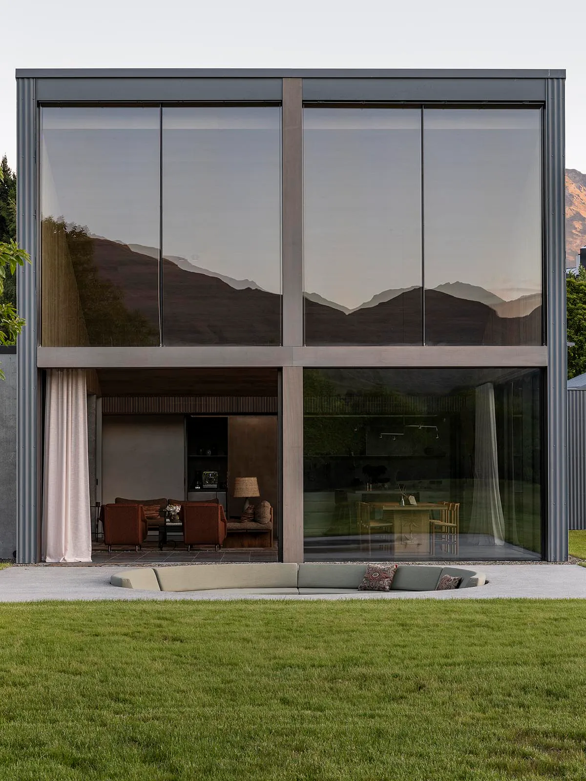 Modern two-story house with floor-to-ceiling glass walls reflecting mountains, open living and dining areas visible, curved outdoor sofa on lawn in foreground.