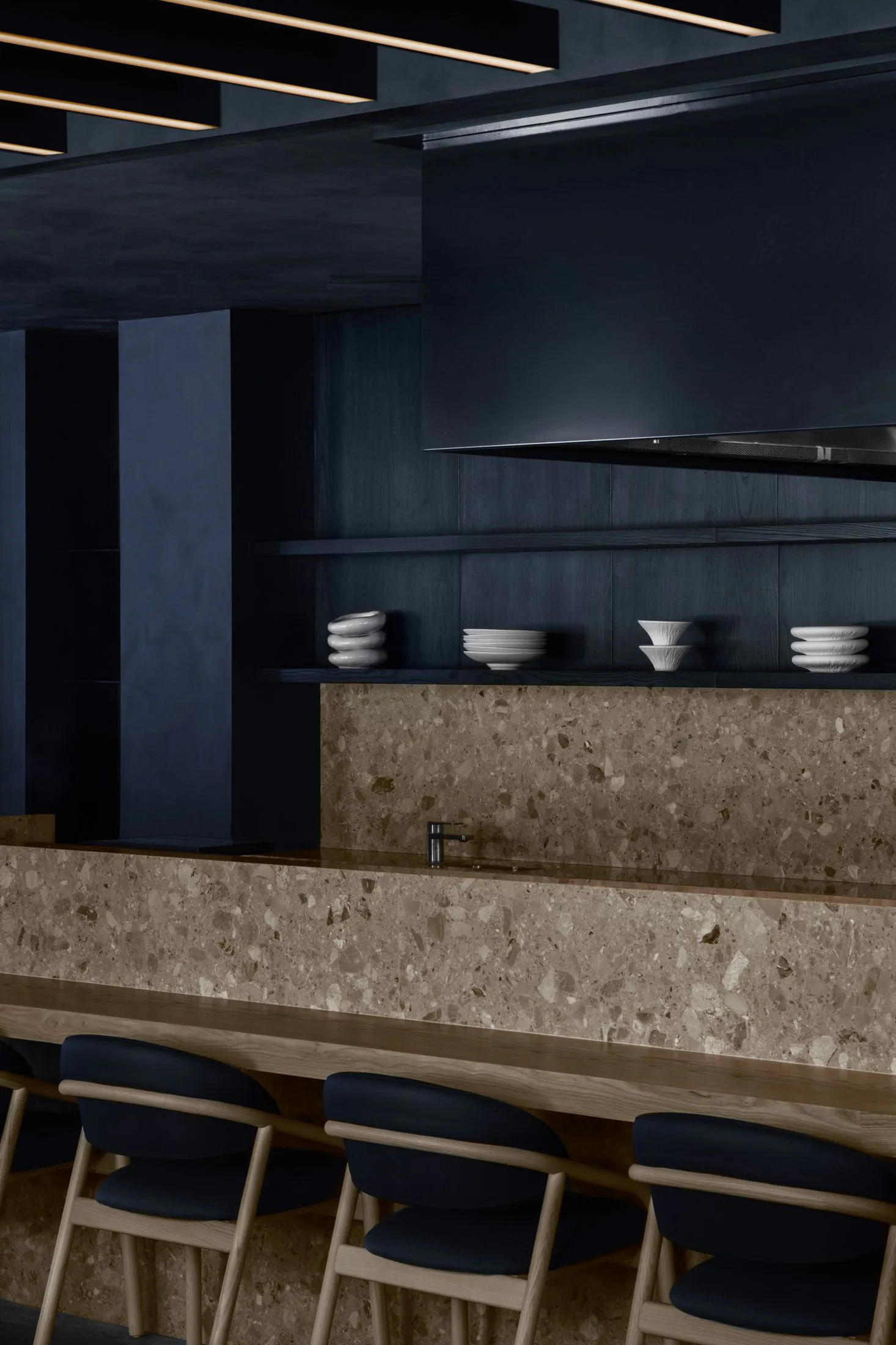 Modern kitchen bar counter featuring beige terrazzo surfaces, dark blue upholstered wooden stools, open shelving with white ceramics, and dramatic linear ceiling lighting.