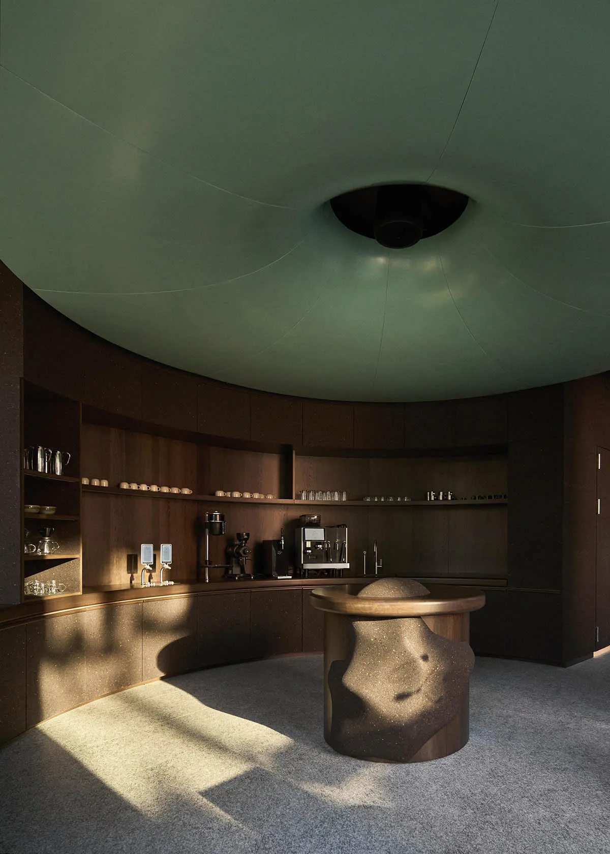 Modern circular coffee bar with dark wood cabinetry, sage green curved ceiling, and a sculptural dark terrazzo central island under dramatic natural light.