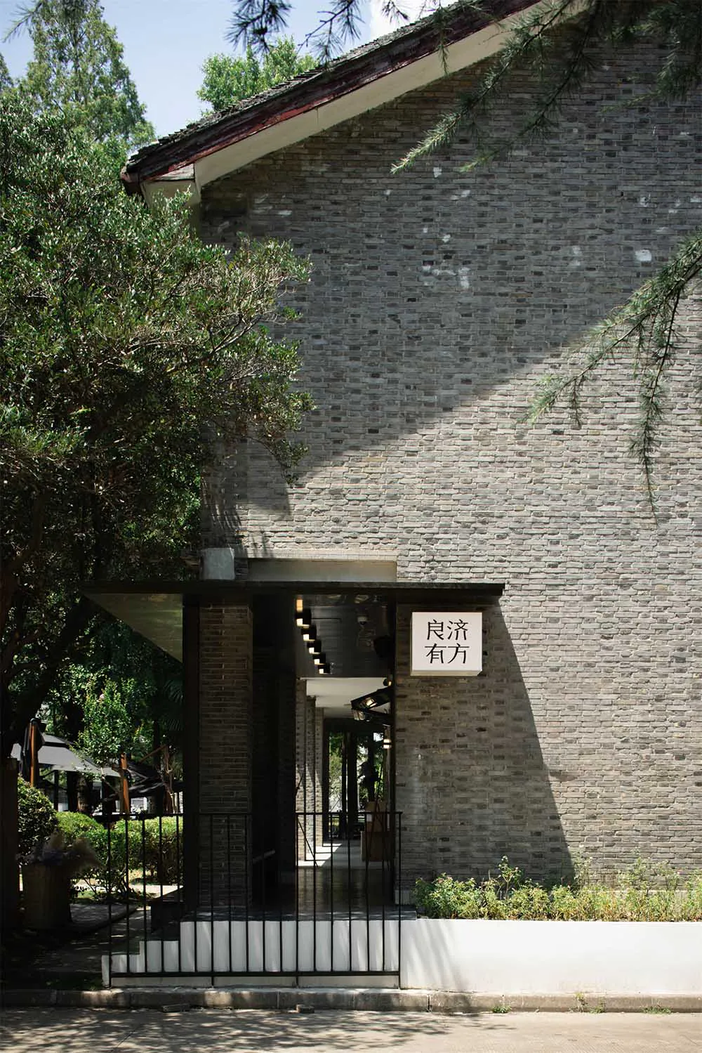 灰磚建築入口外觀,設有現代深色雨棚、黑色金屬柵門、白色階梯,以及帶有中文標誌的白牌,周圍環繞茂密綠樹,陽光充足。