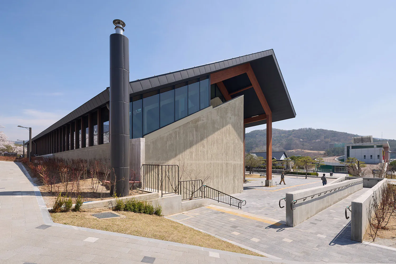 Modern cultural facility featuring exposed concrete walls, large glass windows, a tall cylindrical chimney, and prominent wooden structural canopy, surrounded by paved plazas and hills in the background.