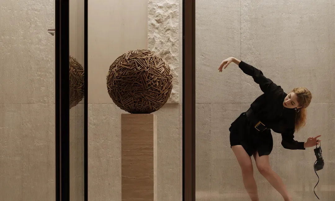 A woman in a black dress posing dynamically next to a large spherical sculpture made of wooden sticks, displayed on a stone pedestal in a minimalist interior with textured walls and large glass panels.