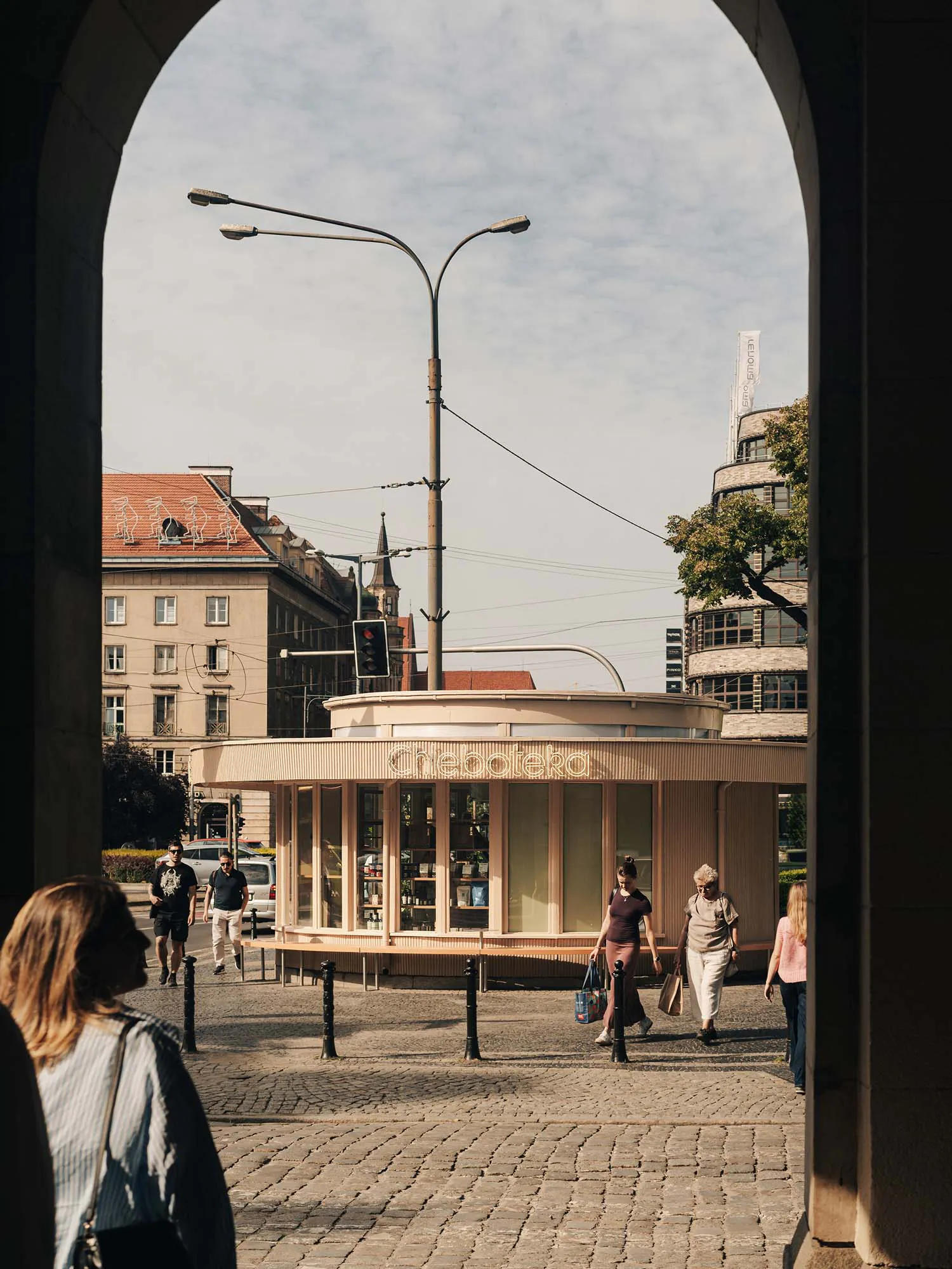 View through a stone arch of a modern curved wooden kiosk named Cheboteka on a sunny cobblestone square in Prague, with pedestrians walking by.