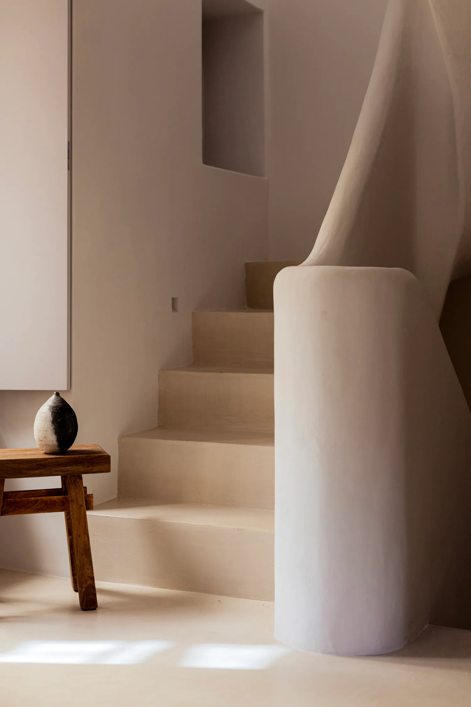 A minimalist interior featuring a curved white plaster staircase, wooden stool with ceramic vase, and soft natural lighting.