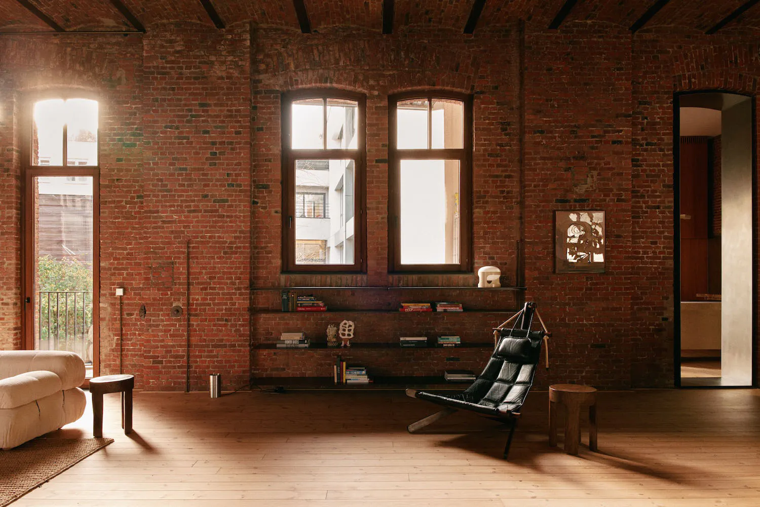Spacious industrial-style living room with red exposed brick walls, wooden floor, large arched windows, black leather sling chair, built-in bookshelves, and a white sofa.