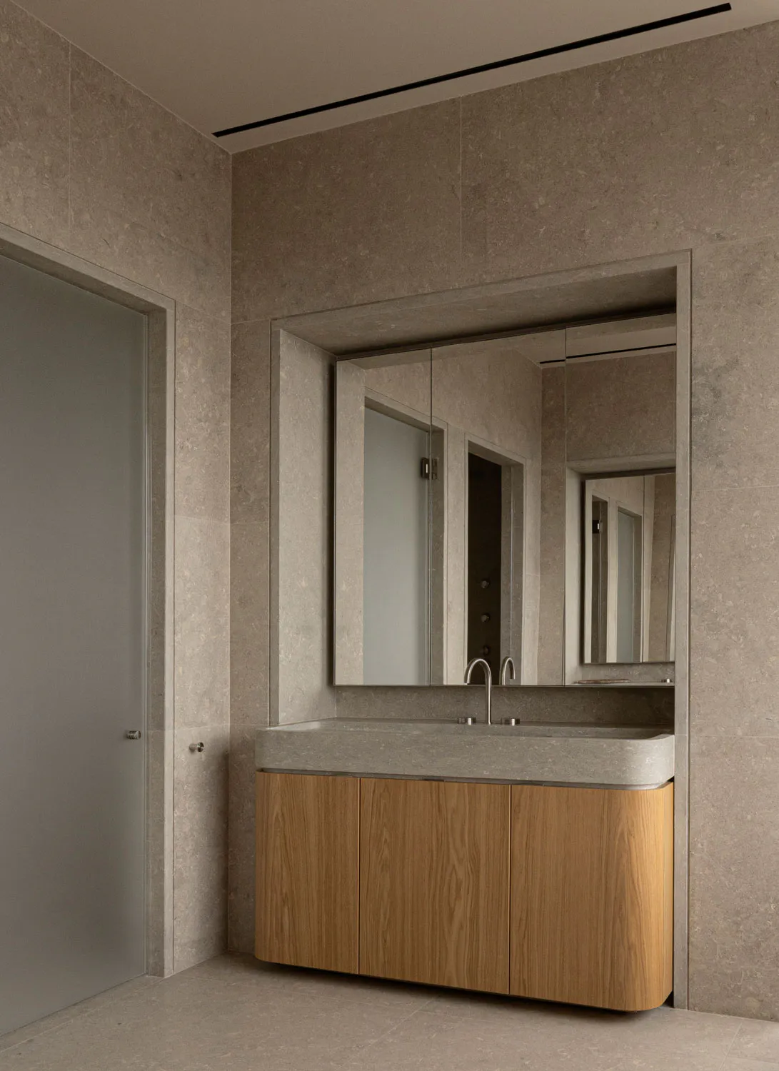 Modern minimalist bathroom corner with large travertine tiled walls, a floating wooden vanity with integrated stone sink and chrome faucet, and a recessed mirrored medicine cabinet reflecting the space.