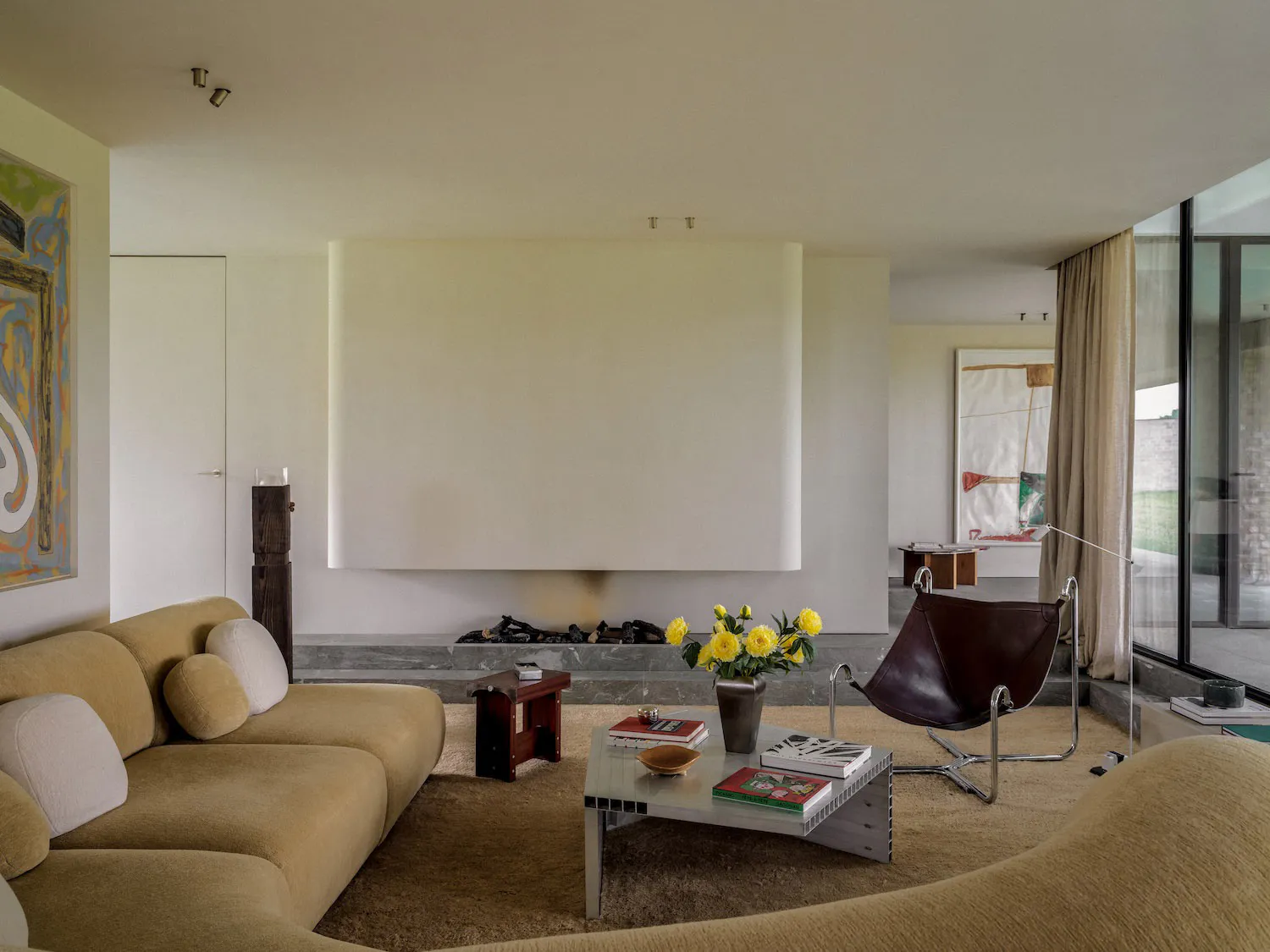Modern minimalist living room featuring a curved beige sectional sofa, dark brown leather butterfly chair, metal and glass coffee table, and a recessed fireplace with large abstract paintings.