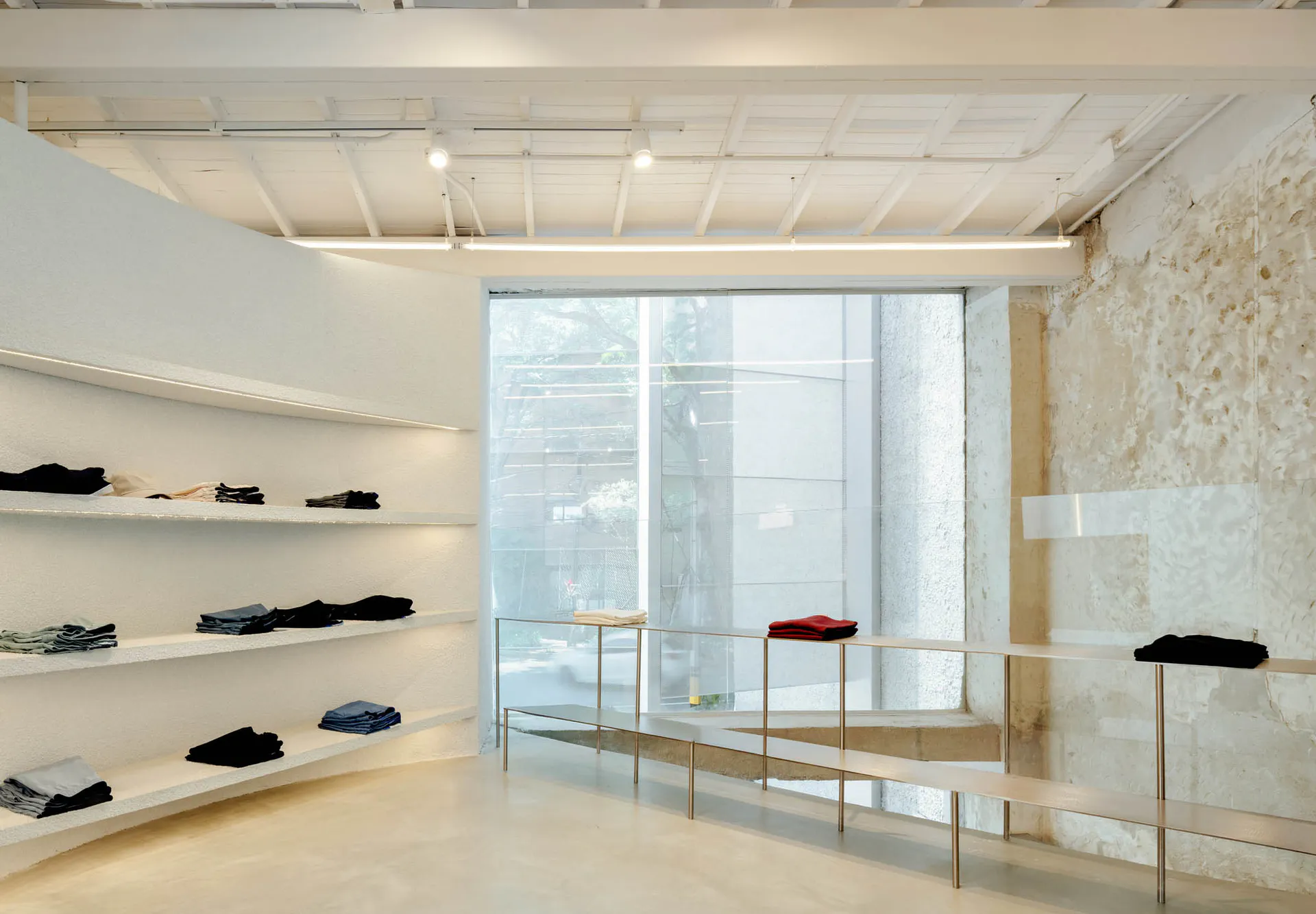 Modern minimalist clothing boutique interior featuring a large exposed concrete wall, white floating display shelves with folded garments, stainless steel clothing racks, and bright natural light from floor-to-ceiling windows.