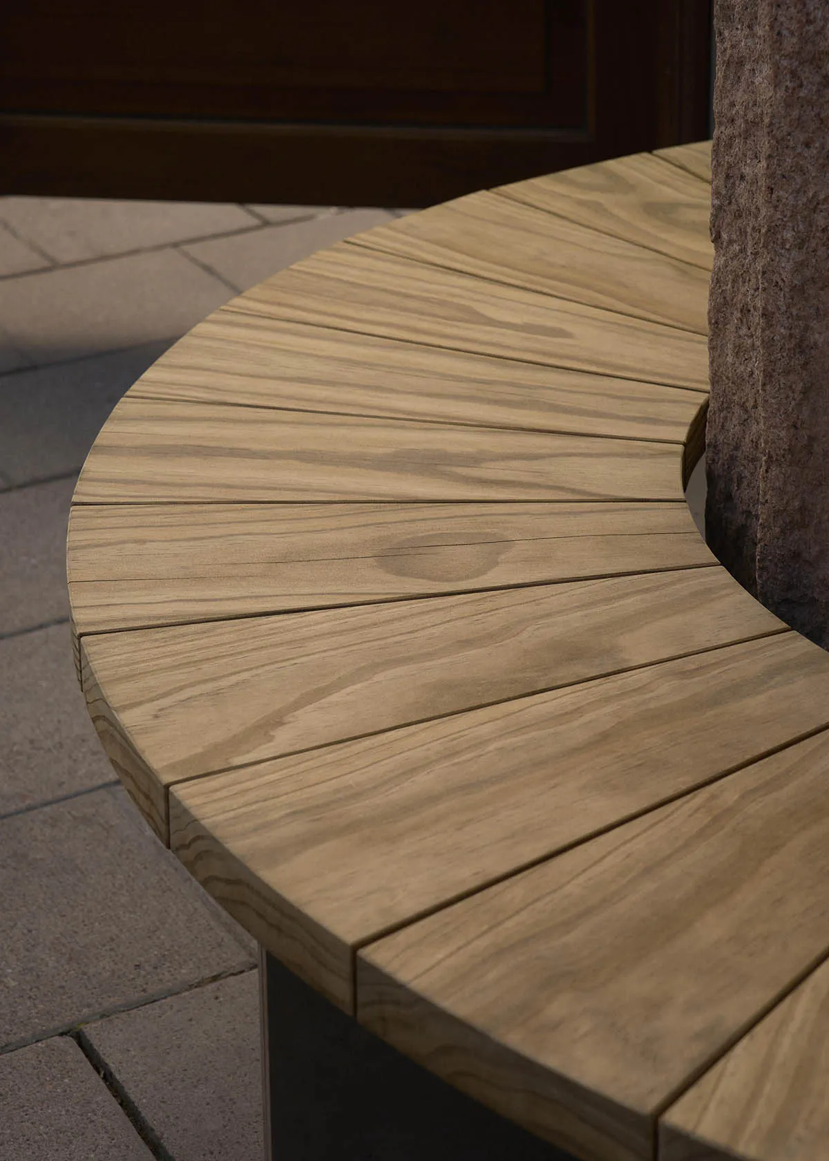 Close-up view of a curved outdoor bench made of light-colored teak wood slats arranged radially around a textured stone pillar, set on a paved stone ground.