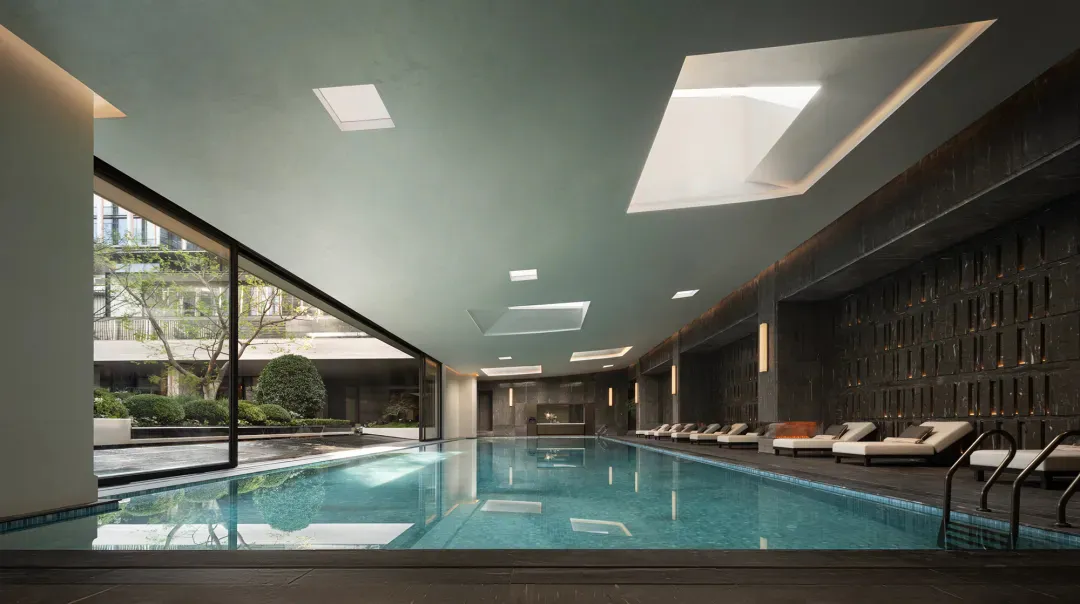 Modern indoor swimming pool in a luxury spa with dark textured stone walls, multiple skylights, and lounge chairs along the side.