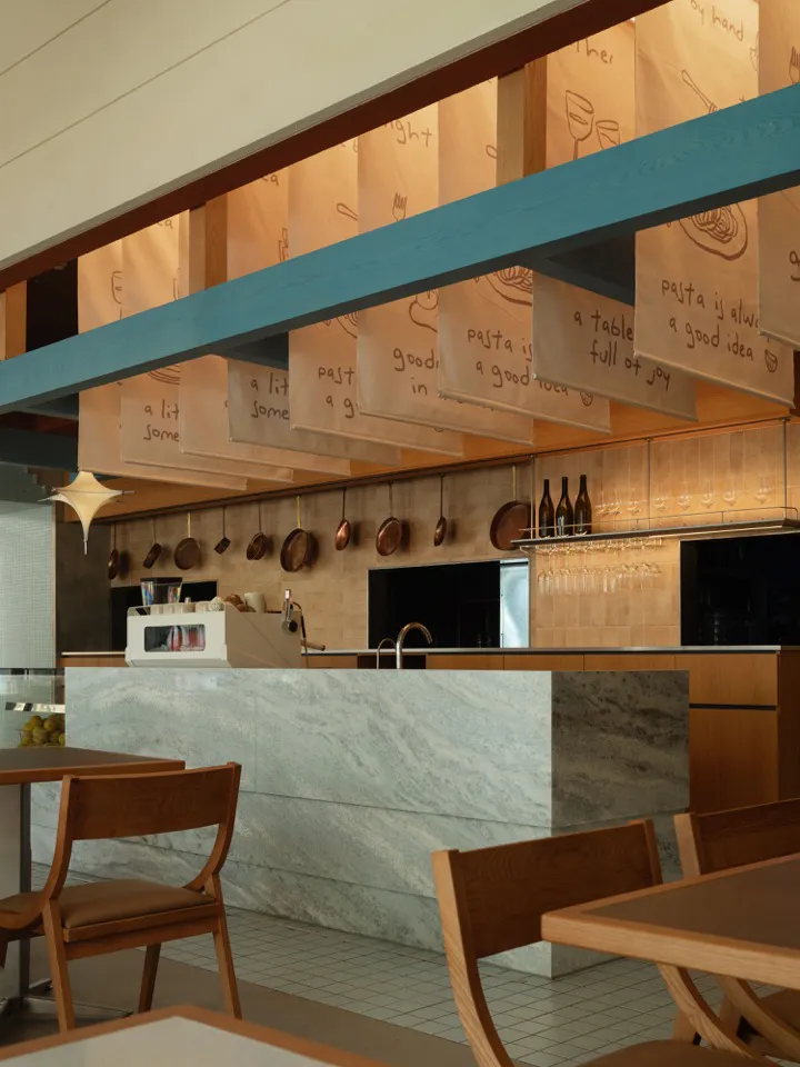 Interior view of a modern Italian restaurant featuring a large marble counter, wooden dining chairs, hanging copper cookware, and fabric banners with handwritten pasta-themed messages suspended from the ceiling.