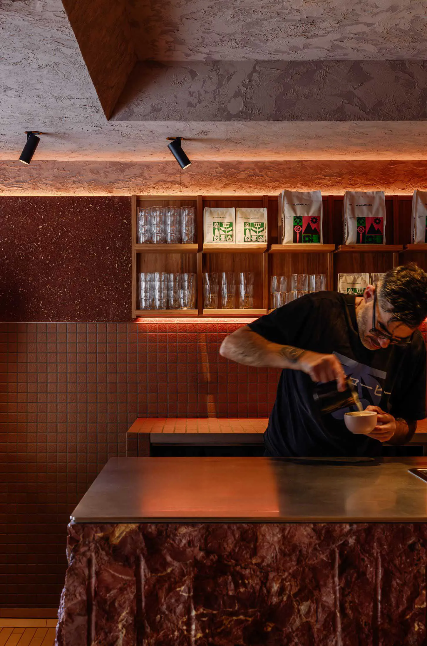 A male barista with glasses and tattoos pouring pour-over coffee behind a marble counter in a modern coffee shop with red tiled walls and wooden display shelves.