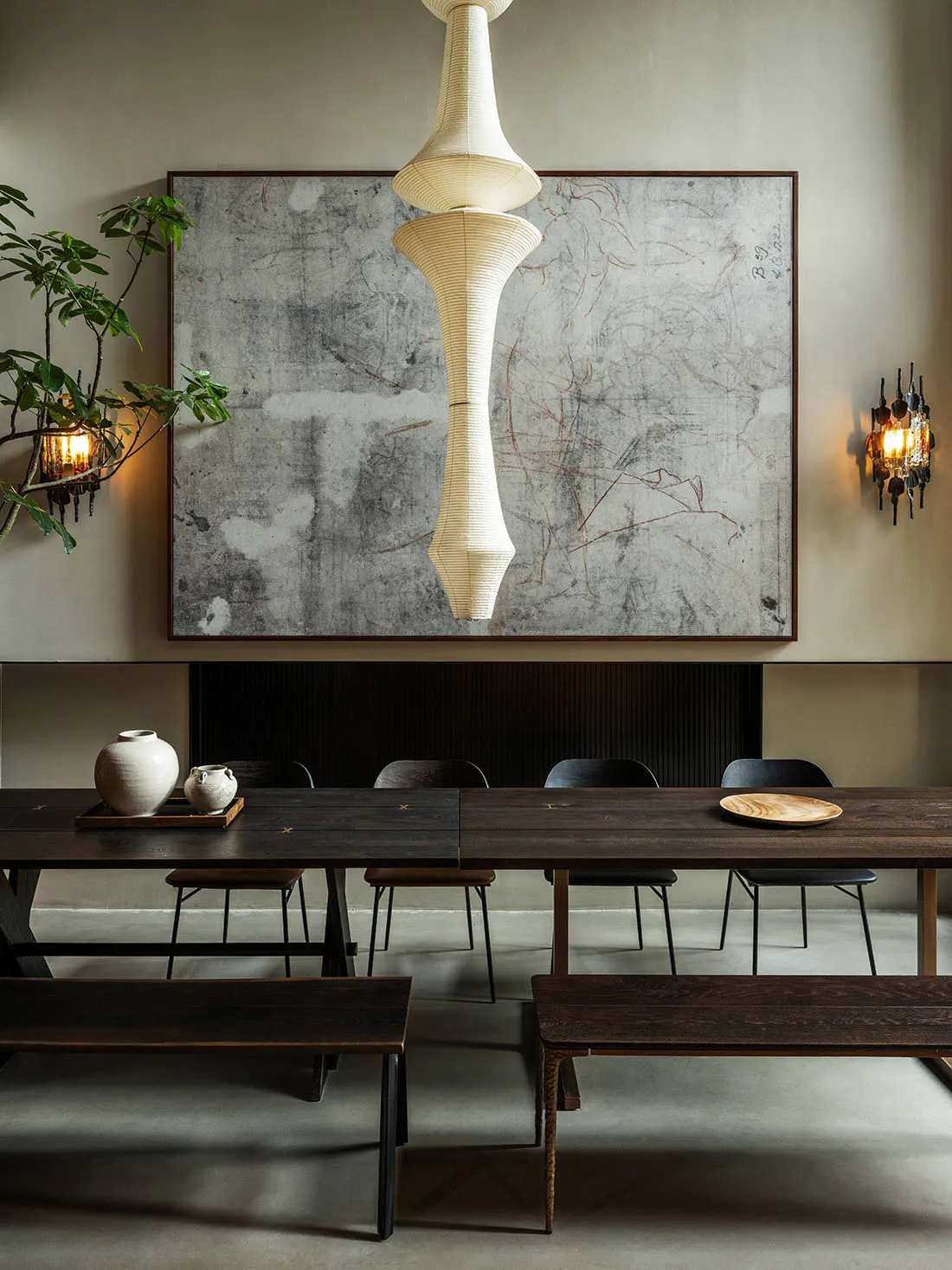 Modern minimalist dining room featuring a long dark wood table, wooden benches, black dining chairs, a large abstract concrete artwork on the wall, a tall sculptural white paper pendant lamp, and warm wall sconces.